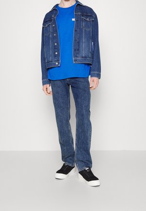 Chaqueta de mezclilla azul con cierre de botones, combinada con una camiseta azul y jeans azul claro. Zapatillas negras completan el conjunto.