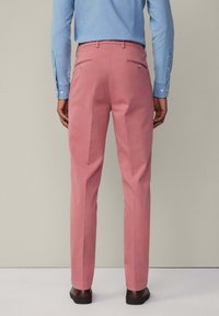 Schmale, pinke Baumwollhosen mit zwei Gesäßtaschen und metallischen Knopfdetails. Kombiniert mit einem blauen Hemd, getragen mit dunklen Schuhen.
