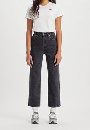 Frau trägt schwarze Jeans mit hoher Taille, weißes Levi's T-Shirt, weiße Sportsocken und graue Turnschuhe, steht vor einem schlichten Hintergrund.