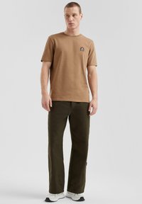 T-shirt in cotone marrone con maniche corte e una piccola etichetta logo, abbinato a pantaloni verde oliva. Il modello indossa sneakers bianche.