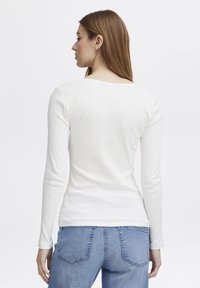 Hvit langermet ribbestrikket topp, tettsittende design, med dyp utringning. Modellen har den på seg med blå denimjeans. Glatt tekstur.