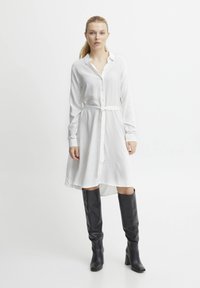 Robe blanche à boutons avec une taille à nouer, manches longues et un ourlet légèrement arrondi. Portée avec des bottes noires en cuir jusqu'aux genoux.