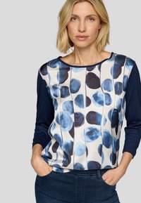 Blau-weiße Bluse mit einem Muster aus kreisförmigen Aquarellen, langen marineblauen Ärmeln und einem runden Ausschnitt. Weicher, glatter Stoff.