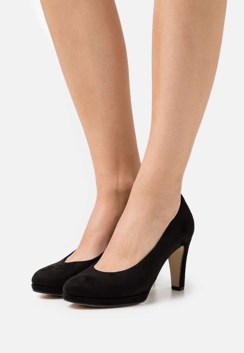 Jambes portant des chaussures à talons noirs fermées, avec un avant arrondi et un talon intérieur beige, posées sur une surface blanche.