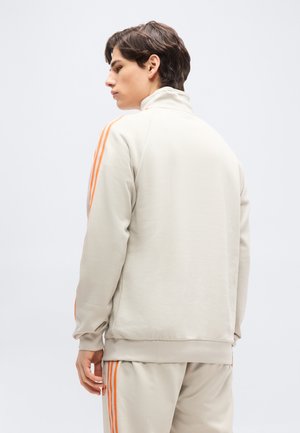 Noor mees külgvaates, seljas beež dressipluus, millel oranžid triibud varrukatel ja pükstel, taustaks on ühtlane sein.