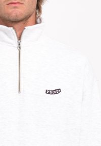 Sweatshirt blanc à fermeture éclair avec un col montant. Présente un petit logo "Volcom" noir sur la poitrine. Texture douce, ressemblant à du polaire.