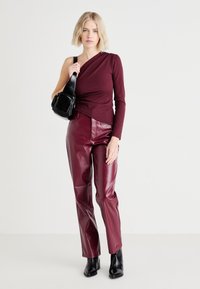 Burgundy top med en axel i kombination med bourgogne-färgade byxor med krokodiltextur. Modellen håller en liten svart handväska och bär svarta ankelskor.