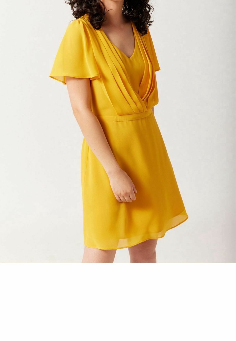 Femme portant une robe courte jaune avec un devant plissé et des manches flottantes, debout devant un fond blanc uni.