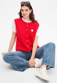 Rote gestrickte Weste mit Knopfverschluss und einem kleinen grünen Etikett, über einem weißen Hemd getragen. Kombiniert mit hellblauen Jeans mit weitem Bein und weißen Turnschuhen.