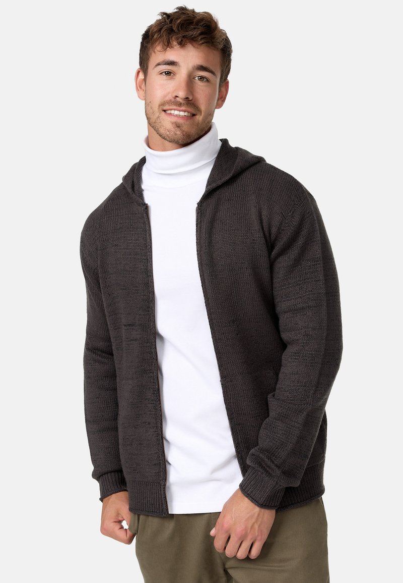 Mörkgrå stickad cardigan med huva, ribbade muddar och dragkedja fram. Bärs över en vit turtleneck. Avslappnad passform och texturerad tyg.