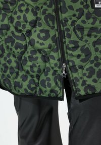 Giacca imbottita verde con stampa leopardata nera, dotata di una zip ornata con un tirante a forma di stella. Pantaloni neri sono parzialmente visibili.