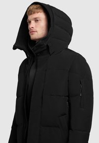 Svart puffjacka med en stor huva, framdragkedja och flera dragkedjefickor. Materialet verkar slätt och isolerat.