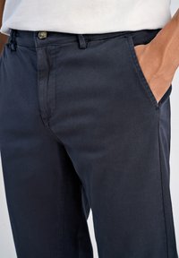 Närbild av en person som bär marinblå byxor med en hand insatt i den högra framfickan, i kombination med en vit skjorta.