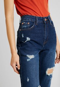 Blå slitna denimjeans med trasiga lappar, hög midja, fem fickor och detaljer i koppar. Strukturerad tyg med synlig slitage.