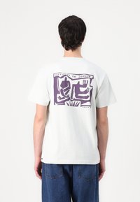 T-shirt en coton blanc avec une illustration graphique violette au dos, représentant des mains et des formes abstraites, avec le texte "REGARDANT LE SOLEIL SE COUCHER."