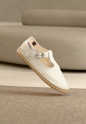 Espadrille en toile blanche avec bride à boucle, semelle tressée et étiquette de taille 37, posée devant un canapé beige et un tapis texturé.