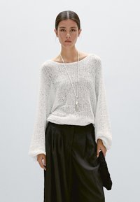 Frau mit zurückgebundenem dunklem Haar trägt einen locker sitzenden weißen Strickpullover, eine lange Kettenhalskette und schwarze High-Waist-Hose, hält eine schwarze, strukturierte Clutch in der Hand.