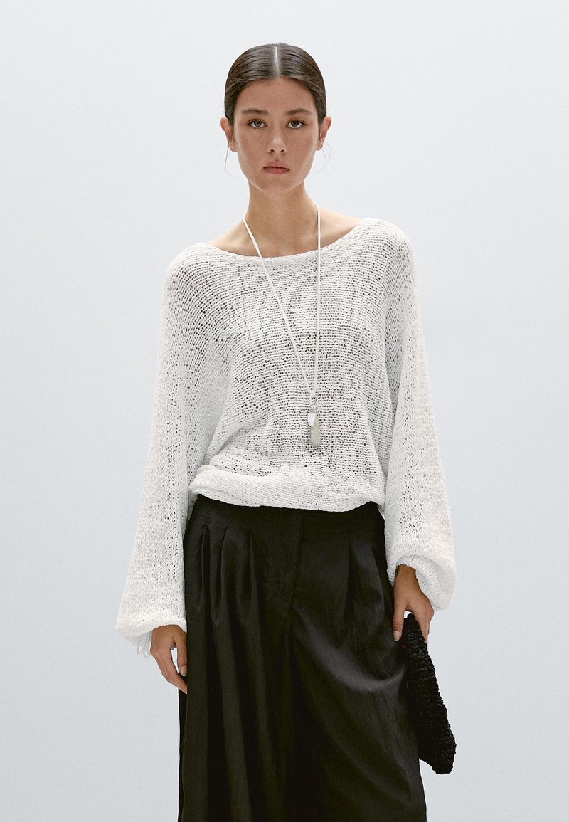 Frau mit zurückgebundenem dunklem Haar trägt einen locker sitzenden weißen Strickpullover, eine lange Kettenhalskette und schwarze High-Waist-Hose, hält eine schwarze, strukturierte Clutch in der Hand.