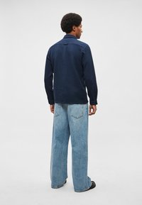 Tengerészkék felgombolható ing texturált mintával, világoskék bő szárú farmerrel párosítva. Az ing kis gallérral és gombokkal ellátott mandzsettával rendelkezik.