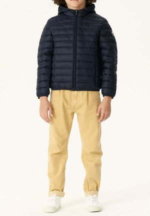 Giacca piumino blu navy con cuciture orizzontali e cappuccio, abbinata a pantaloni beige chiaro e scarpe bianche. Design semplice, materiale leggero.