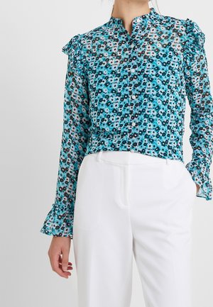 Blouse boutonnée à manches longues avec motif floral bleu et épaules à volants, associée à un pantalon blanc taille haute.