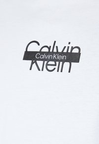 Camiseta blanca de algodón con un logo gráfico negro de "Calvin Klein" en una fuente moderna con letras superpuestas.