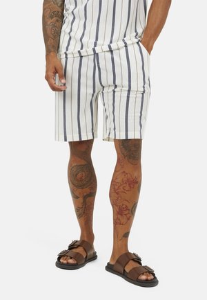 Mężczyzna w białych krótkich spodenkach w pionowe paski i brązowych sandałach, pokazujący tatuaże na ramionach i nogach na jednolitym tle.