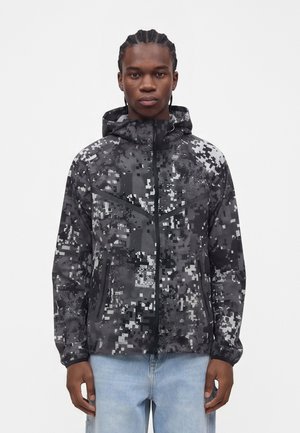 Mand iført en sort og grå pixeleret camouflage hættetrøje med lynlås, stående foran en ensfarvet lys baggrund.