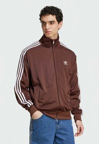 Bruine ritsjas van gladde stof, met witte drie-strepen accenten op de mouwen en een klein wit logo op de borst.