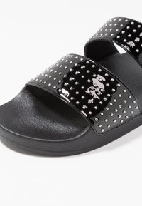 Sandales à glissière noires avec deux brides brillantes ornées de clous en argent. Semelle noire texturée offrant adhérence et confort.