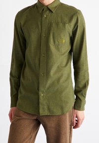 Camisa verde de manga larga en una tela texturizada, con cuello tipo polo abotonado, un bolsillo en el pecho y un pequeño logo en el bolsillo.