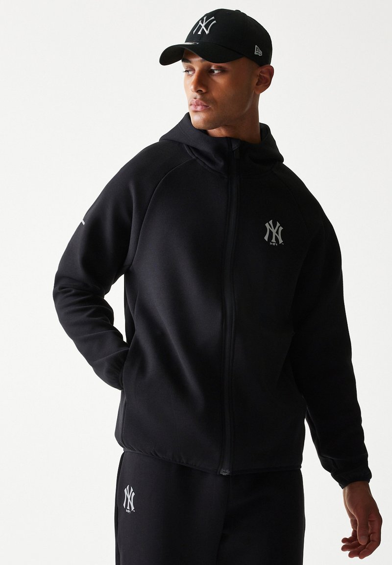 Άνδρας που φοράει μαύρο καπέλο New York Yankees, μαύρο φούτερ με φερμουάρ και μαύρο παντελόνι με λογότυπα Yankees στο στήθος και το πόδι.