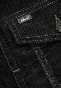 Schwarzer Denim-Stoff mit einer sichtbaren Tasche, die einen silbernen Knopf und ein gesticktes Etikett mit der Aufschrift "Reell" in weißer Schrift aufweist.