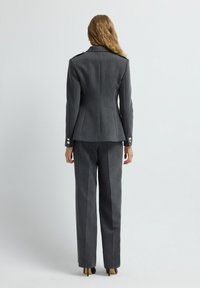 Traje gris a medida que presenta un blazer ajustado con acentos en los hombros y pantalones rectos de cintura alta, ambos en un tejido texturizado.