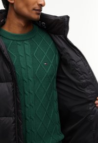 Homme portant un pull vert en maille torsadée avec un petit logo et une doudoune noire ouverte pour montrer le pull, regardant vers la gauche.
