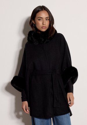 Femme portant un manteau noir ceinturé avec une bordure en fourrure au col et aux poignets, associé à un jean bleu, debout devant un fond uni.