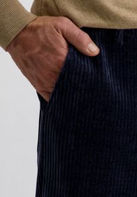Volwassen hand rustend in de voorzak van donkerblauwe corduroybroek, met een beige trui met lange mouwen.