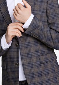 Giacca da completo a quadri marrone con dettagli blu, dotata di due bottoni, tasche a filetto e una camicia bianca liscia sotto.