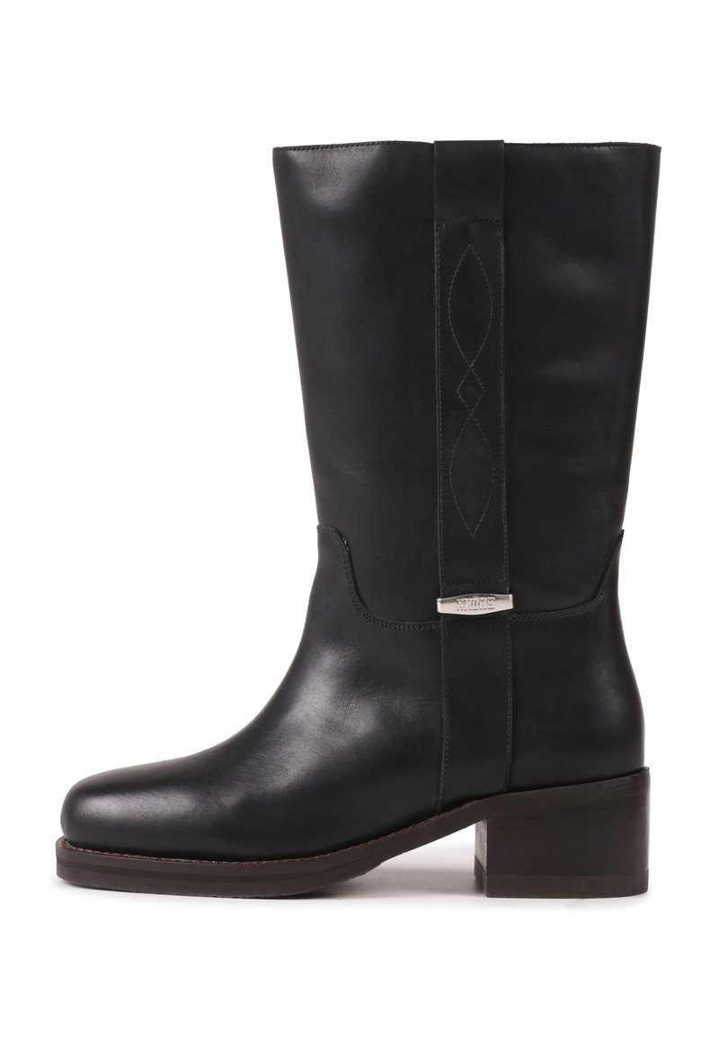 Μαύρη δερμάτινη μπότα μήκους ως τη μέση της γάμπας με χαμηλό τετράγωνο τακούνι, στρογγυλεμένη μύτη, διακοσμητικό ράψιμο στον άξονα και μεταλλική λεπτομέρεια στο πλαϊνό λουρί.