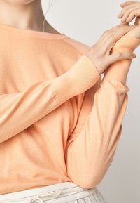 Jasnopomarańczowy sweter dziergany z długimi rękawami, o luźnym kroju i ściągaczach na rękawach. Miękka tekstura i minimalistyczny design.