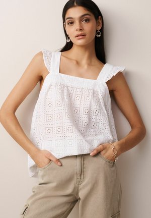 Hvid bomulds tanktop med firkantet udskæring og flæse-stropper, prydet med indviklede hulmønstre. Sammen med beige bukser, der har lommer.