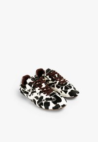 Zapatillas con estampado de vaca en blanco y negro, cordones marrones, material de ante, forma redondeada y superficie texturizada. Detalles plateados en el talón.
