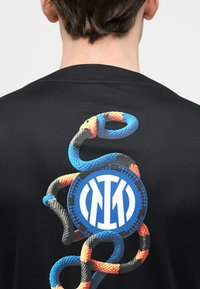 Πίσω μέρος μαύρου πουκάμισου με σχέδιο σχοινιού σε μπλε, πορτοκαλί και μαύρο που περιβάλλει ένα κυκλικό μπλε και λευκό λογότυπο με περιπλεγμένα σχέδια.