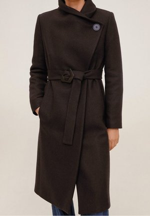 Kobieta w ciemnobrązowym wełnianym płaszczu z paskiem, z asymetrycznym kołnierzem zapinanym na guziki i kieszeniami z boku, w połączeniu z niebieskimi dżinsami.