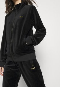 Žena v černém velurovém sportovním obleku FILA s bundou na zip a odpovídajícími kalhotami, stojící na jednoduchém světlém pozadí.