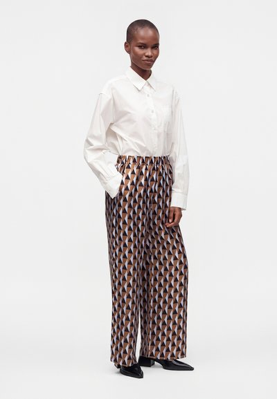 Personne portant une chemise blanche boutonnée et un pantalon à jambes larges avec un motif triangulaire géométrique marron, noir et blanc, debout avec une main dans la poche.