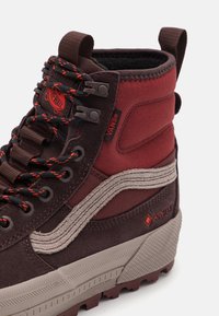 Visoke smeđe i crvene Vans čizme, izrađene od osnovnog materijala od antilopa i tkanine, sa teksturiranim sivim potplatom, narančastim vezicama i GORE-TEX brendiranim detaljima.