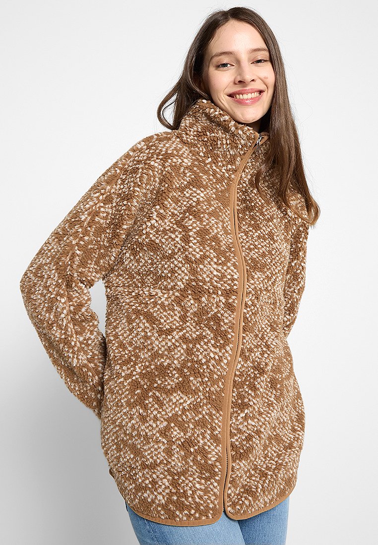 Femme souriante portant une veste en polaire zippée texturée marron et blanche avec un col haut et un jean bleu clair, contre un fond uni.