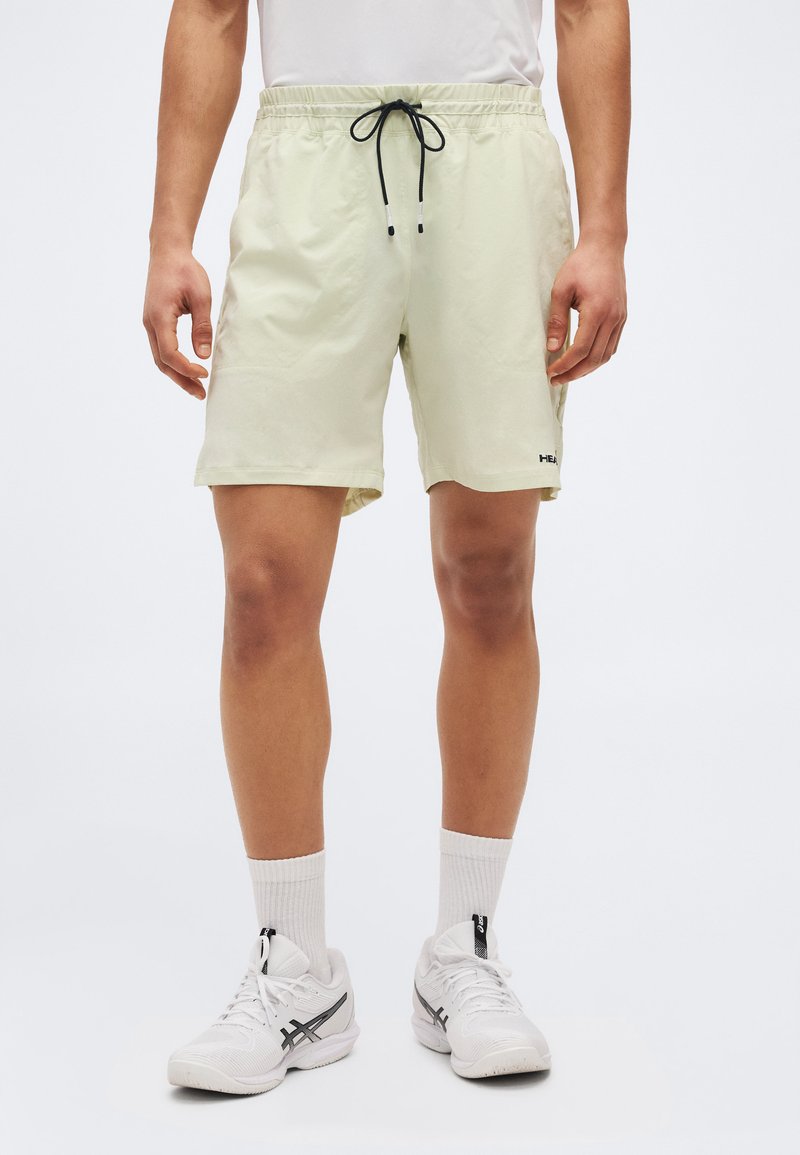 Άνδρας που φοράει ανοιχτό μπεζ σορτς με κορδόνι, λευκές κάλτσες πλήρους μήκους και λευκά παπούτσια τρεξίματος Asics, στέκεται μπροστά σε απλό λευκό φόντο.