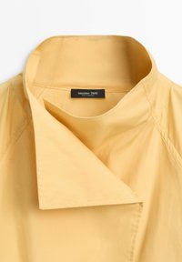 Beige Hemd mit hohem Kragen, breitem, gefaltetem Kragen und sichtbar schwarzem „Massimo Dutti“-Etikett im Halsausschnitt.